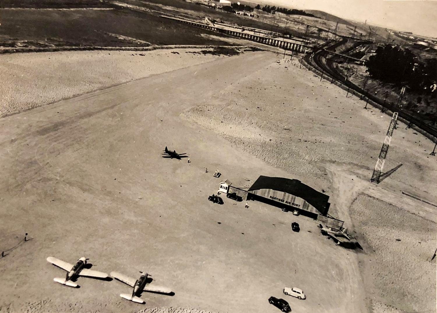 Aerial view of Martinez Airport: a dirt strip on filled ground beside railroad tracks, a hangar lettered MARTINEZ AIRPORT, light aircraft and cars