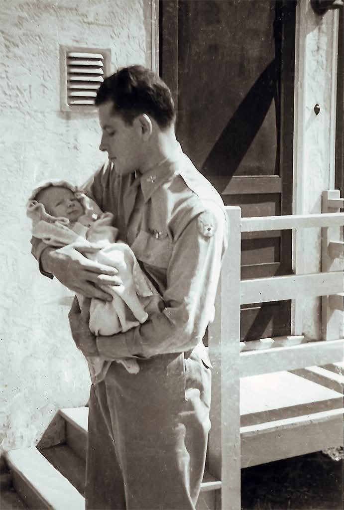 Frank Calicura in khaki on a porch, holding his infant son Jimmy against a stucco wall and a white railing