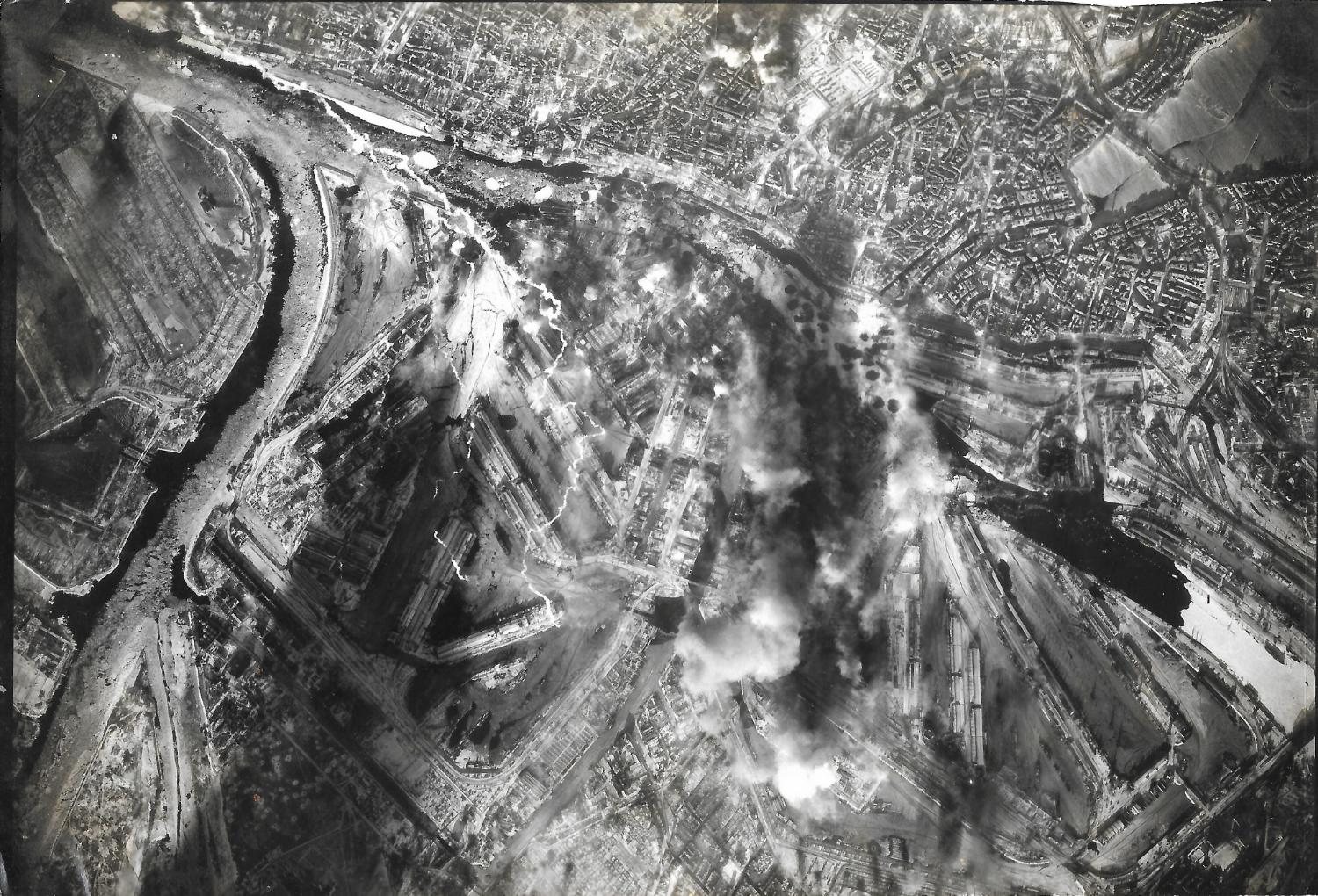 Vertical aerial photograph of a port city, smoke rising from docks and rail yards along a river