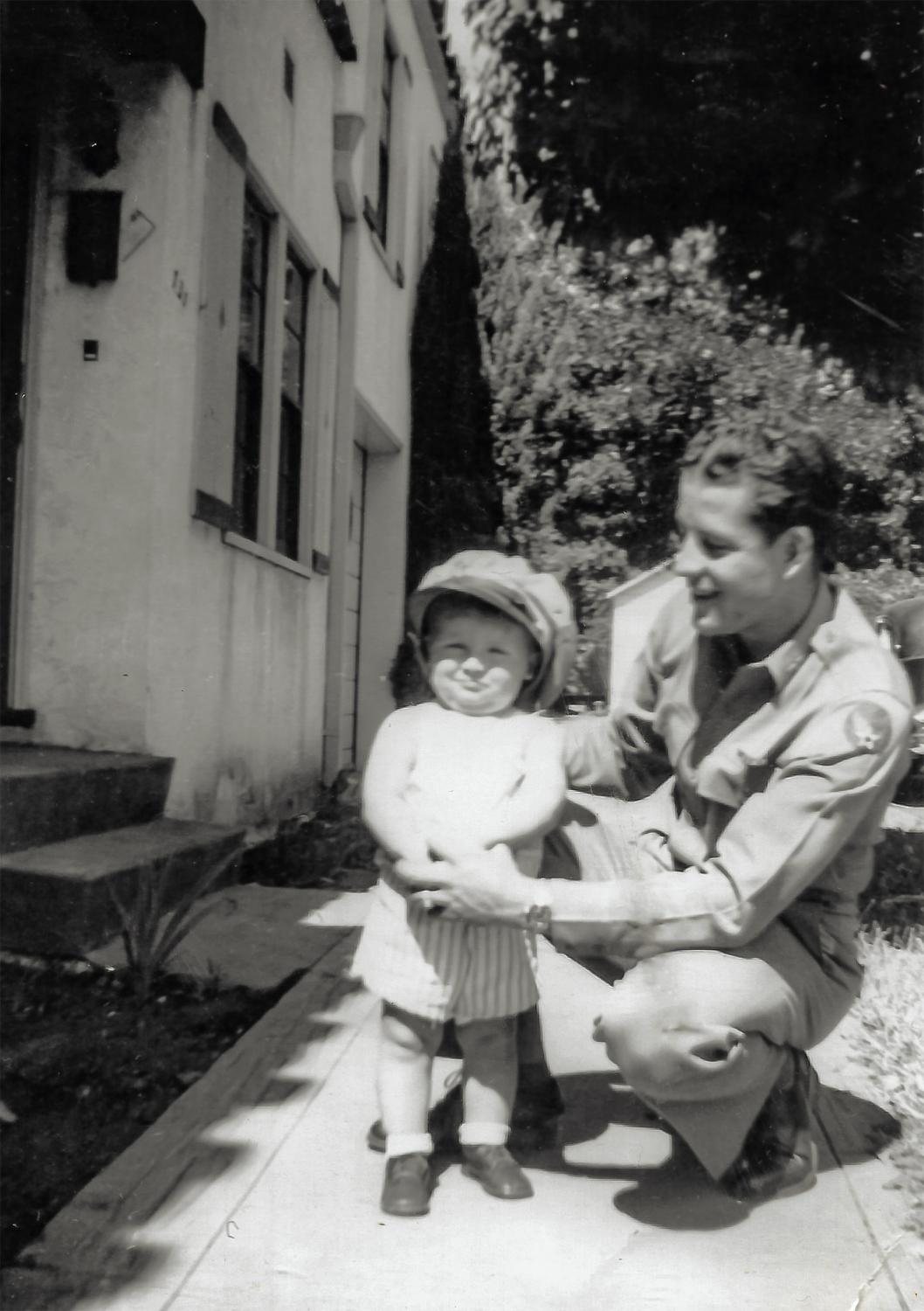 Frank Calicura in khaki, Eighth Air Force patch on the shoulder, kneeling on a sidewalk with a toddler in a hat and striped shorts