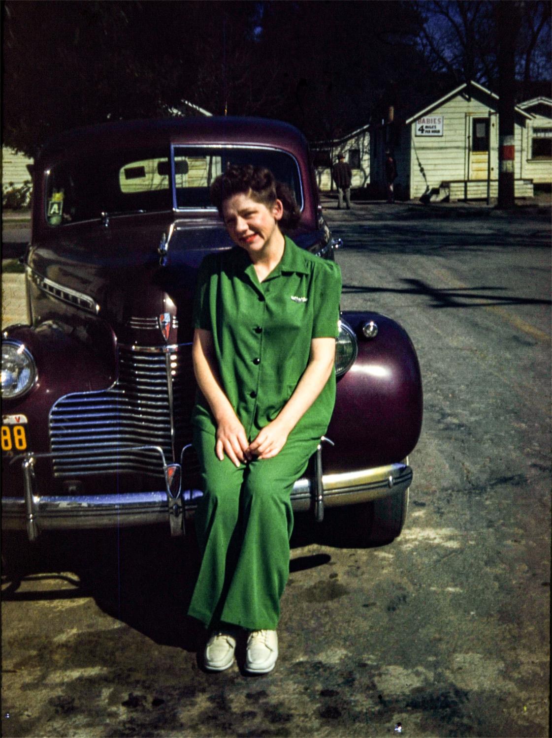 Joyce Calicura in a green dress sitting on the bumper of a maroon car in front of 731 Mellus Street, 1943